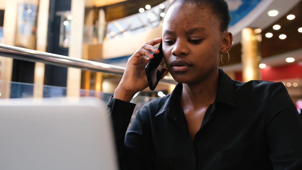 An African woman works at a computer and talks on the phone while typing on the keyboard.