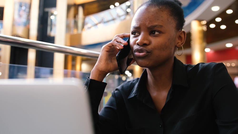 An African woman works at a computer and talks on the phone while typing on the keyboard.