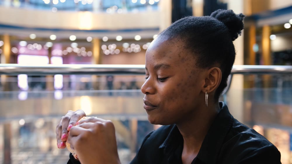 Young african american woman eating fast food in a restaurant enjoying delicious juicy burger