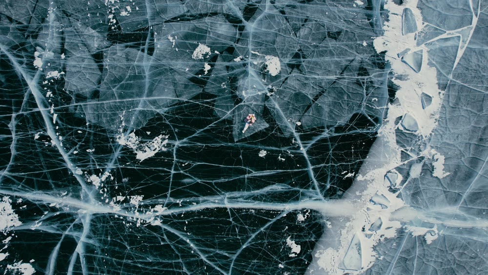 A tourist girl in bright clothes and sunglasses smiles happily, lies on the frozen blue ice of Lake Baikal and controls the drone up. An incredible pattern of crack bubbles. Siberia Russia tourist attraction.