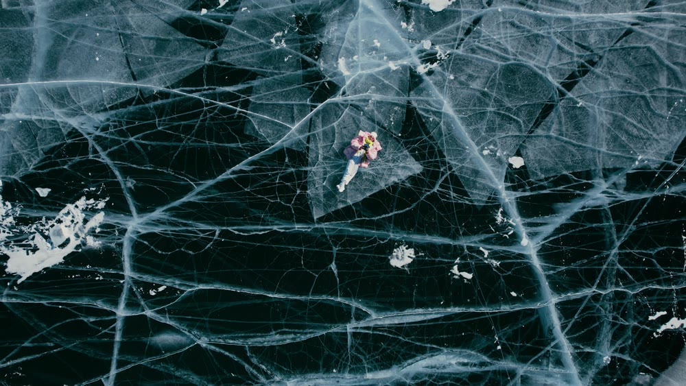 A tourist girl in bright clothes and sunglasses smiles happily, lies on the frozen blue ice of Lake Baikal and controls the drone up. An incredible pattern of crack bubbles. Siberia Russia tourist attraction.