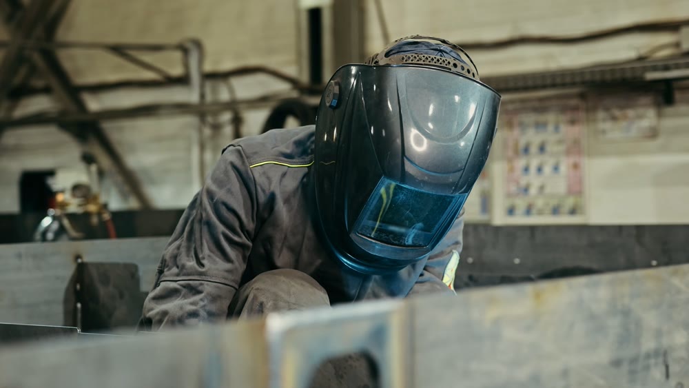 Employee in safety helmet carefully working on steel seam with welding torch amidst heavy machinery